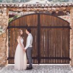 A man and woman dressed for their wedding and holding hands in front of the gate of a villa