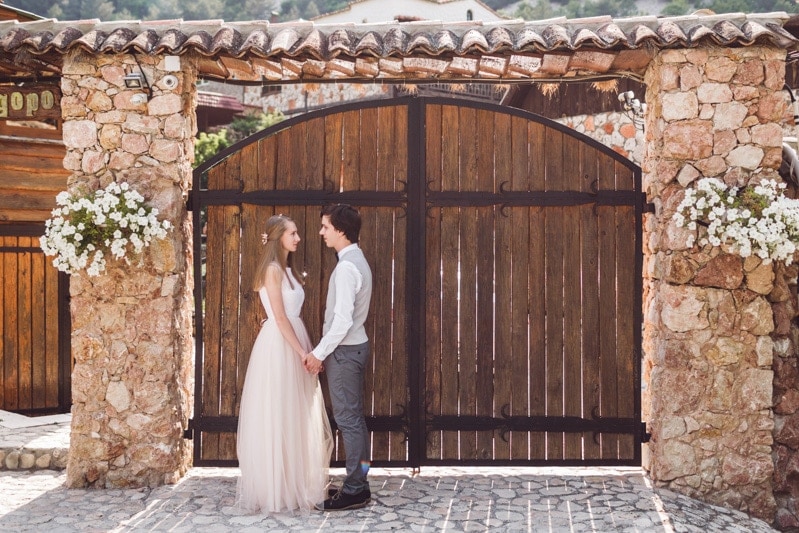 A man and woman dressed for their wedding and holding hands in front of the gate of a villa